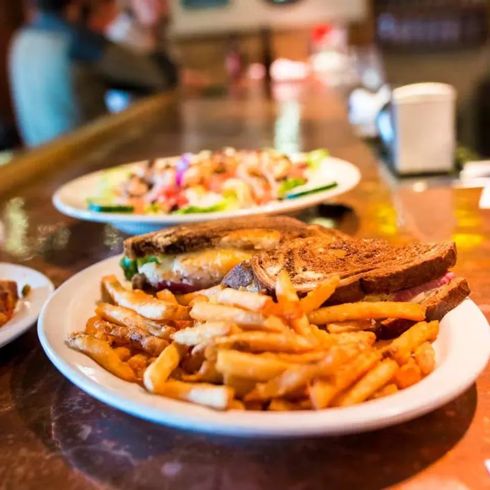 Plate with French fries, grilled sandwich, and sliced steak. In the background, a salad with vegetables and dressing, on a wooden table in a restaurant setting.