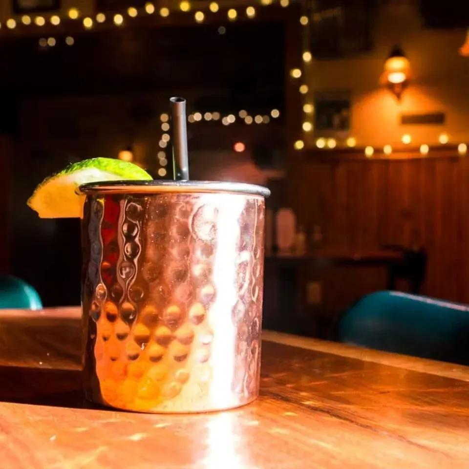 A copper cocktail shaker with a lime wedge on the rim and a black straw, placed on a wooden bar counter with a blurred background of a bar or restaurant interior.